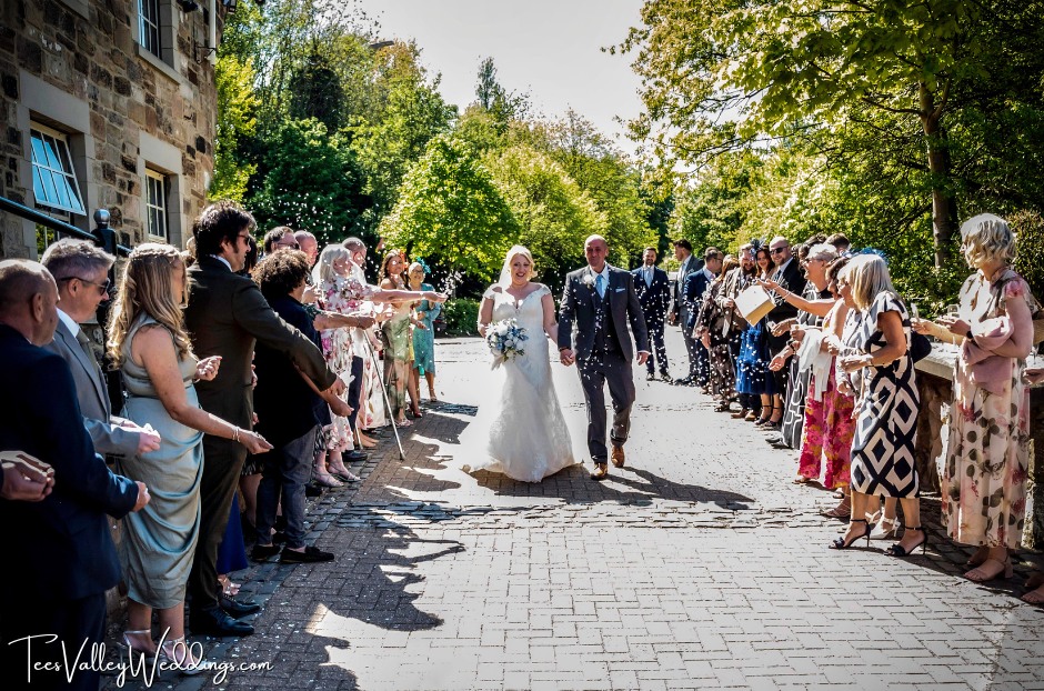 A joyful moment as the newlyweds step out of Webster’s Ropery in Sunderland, walking hand in hand down the center of a crowd. Friends and family surround them, tossing colorful confetti into the air while cheering and shouting congratulations. The couple beams with happiness, their wedding attire shimmering in the evening light.