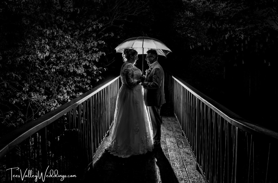 A newlywed couple stands outside the Tree Houses at Ramside Hall late at night, illuminated by off-camera flash. They hold an umbrella, creating a striking silhouette against the darkness. This dramatic signature image by the wedding photographer captures their moment beautifully.