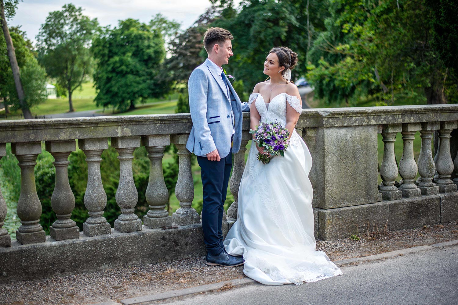 Wedding couple standing together by the stone balustrade at Wynyard Hall, holding a bouquet of purple and white flowers.