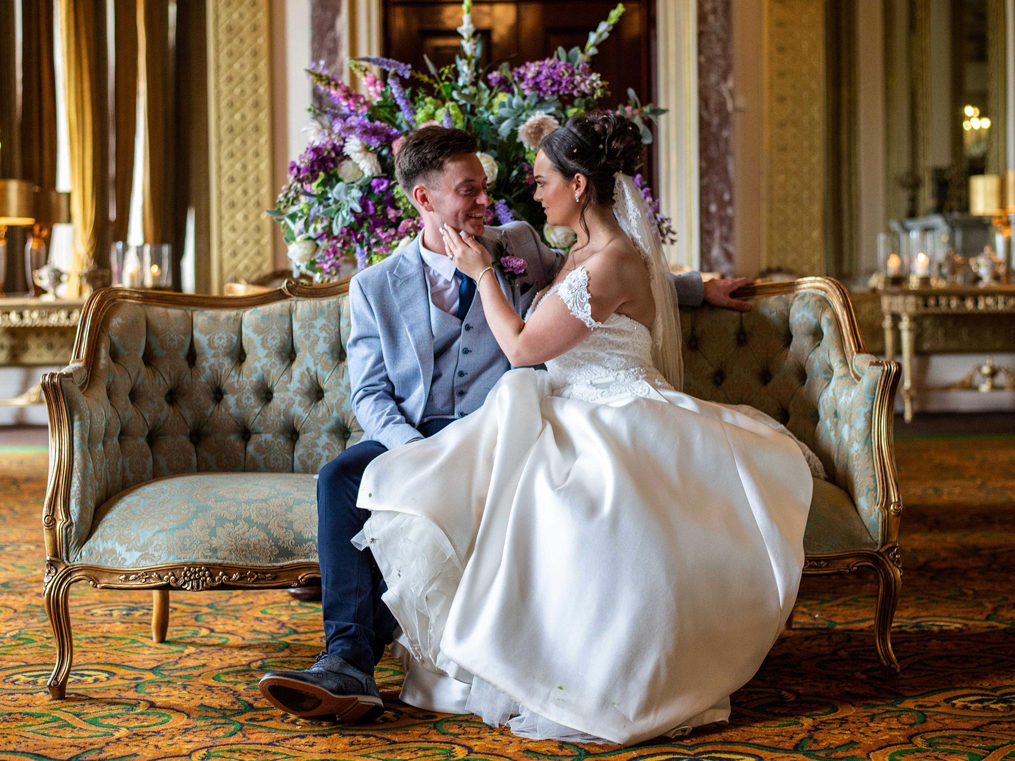 Wedding couple seated on an ornate sofa in the State Room at Wynyard Hall, surrounded by elegant décor and floral arrangements.